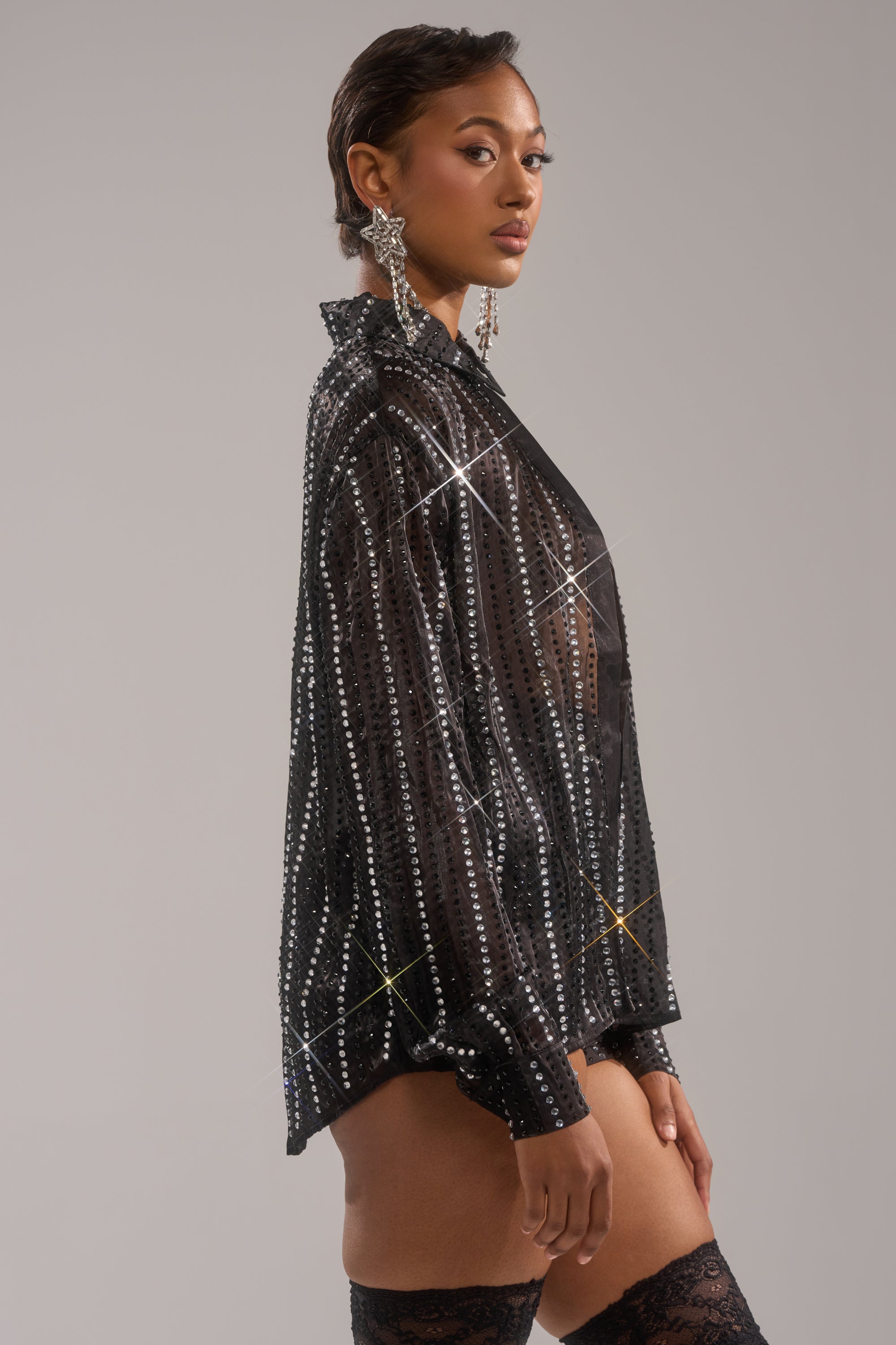 A woman poses in profile wearing the DANCE THE NIGHT AWAY BUTTON DOWN blouse, statement earrings, and lace thigh-high stockings against a plain light gray background.