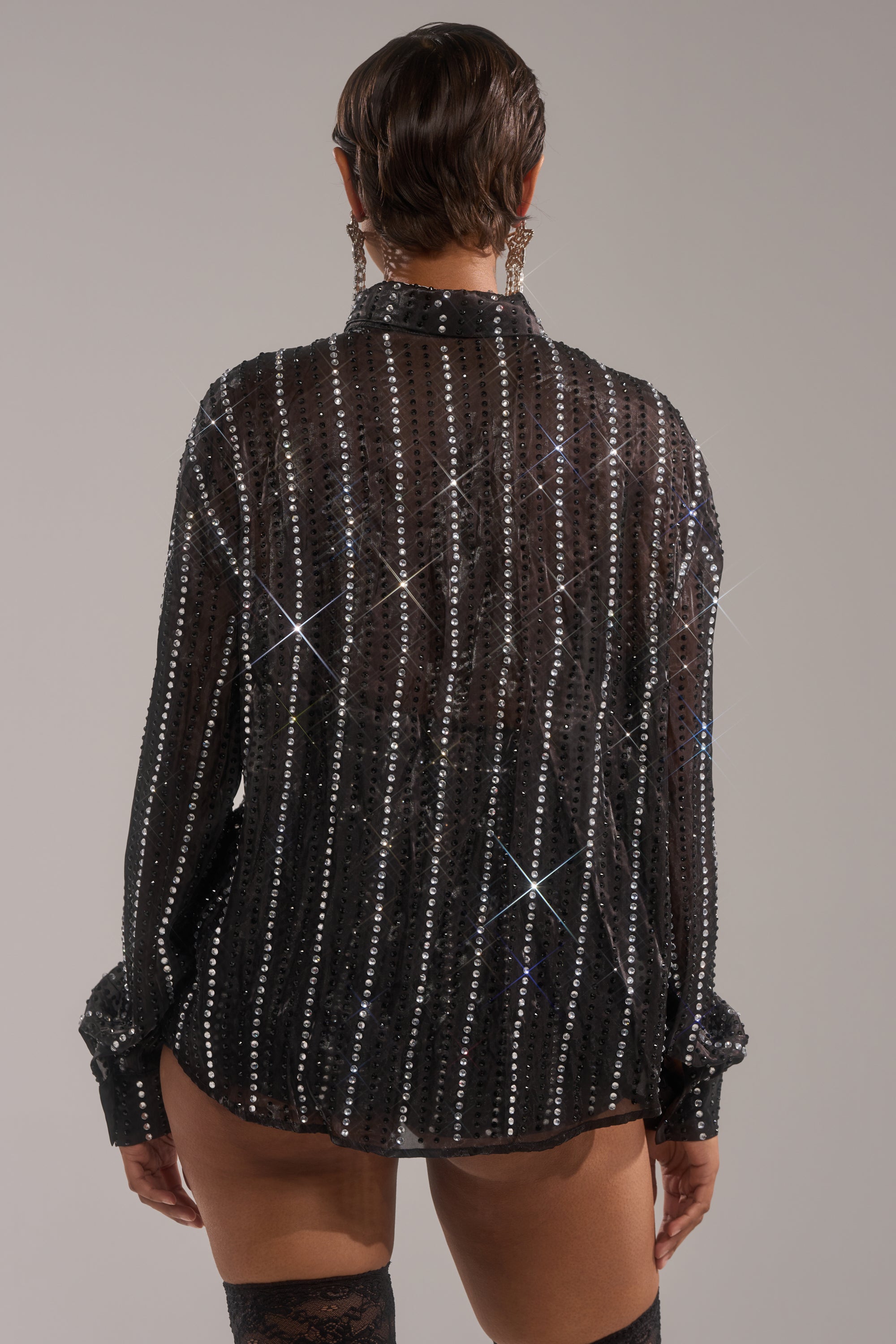 Person with short, slicked-back hair, wearing the DANCE THE NIGHT AWAY BUTTON DOWN—a sheer chiffon top with glittery vertical stripes and earrings—shown from the back. The see-through shirt reveals some skin underneath.