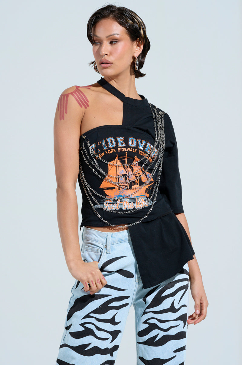 With short dark hair, someone wears the FEEL THE VIBE TEE, a black top with an orange graphic design. They pair it with light blue jeans featuring black zebra stripes and accessorize with faux silver chain links, standing confidently while looking slightly to the side.