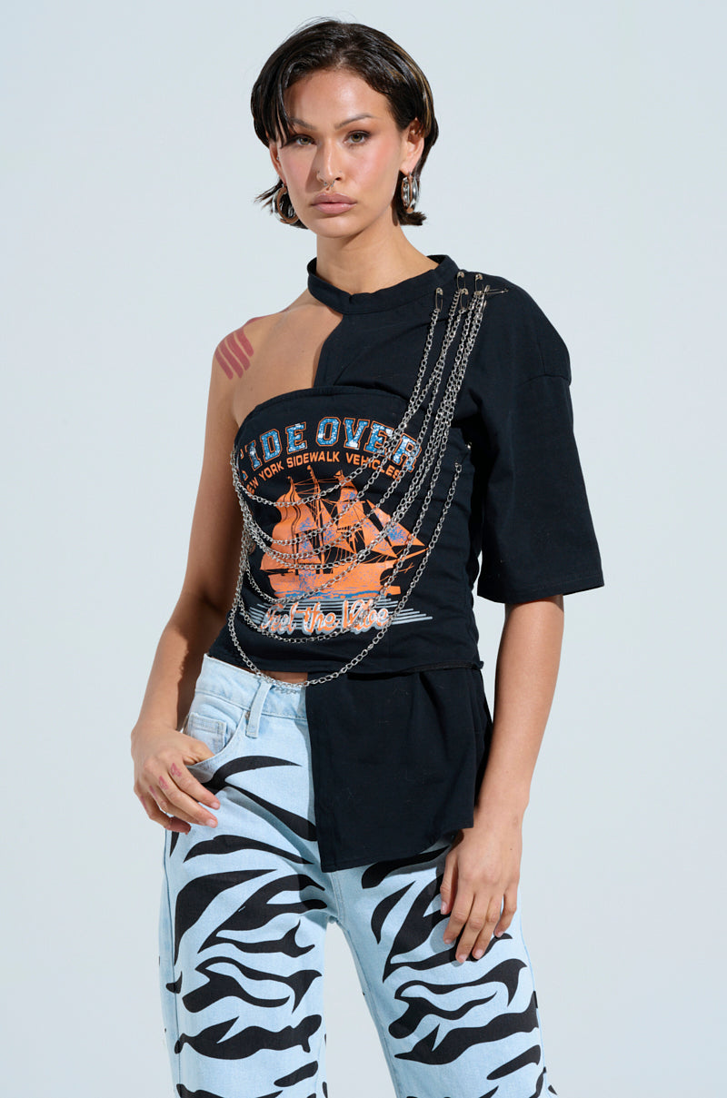 A person confidently wears the FEEL THE VIBE TEE, featuring a bold graphic and faux silver metallic chains. They're paired with light blue pants adorned with striking black zebra stripes on a solid light gray background.