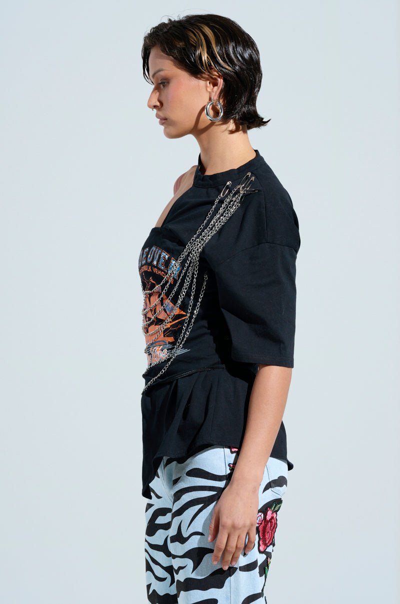 A person with short dark hair stands in profile against a plain background, wearing the FEEL THE VIBE TEE with faux silver metallic chain links. They pair it with light blue pants featuring zebra stripes and red rose patterns, completed by silver hoop earrings for a striking look.