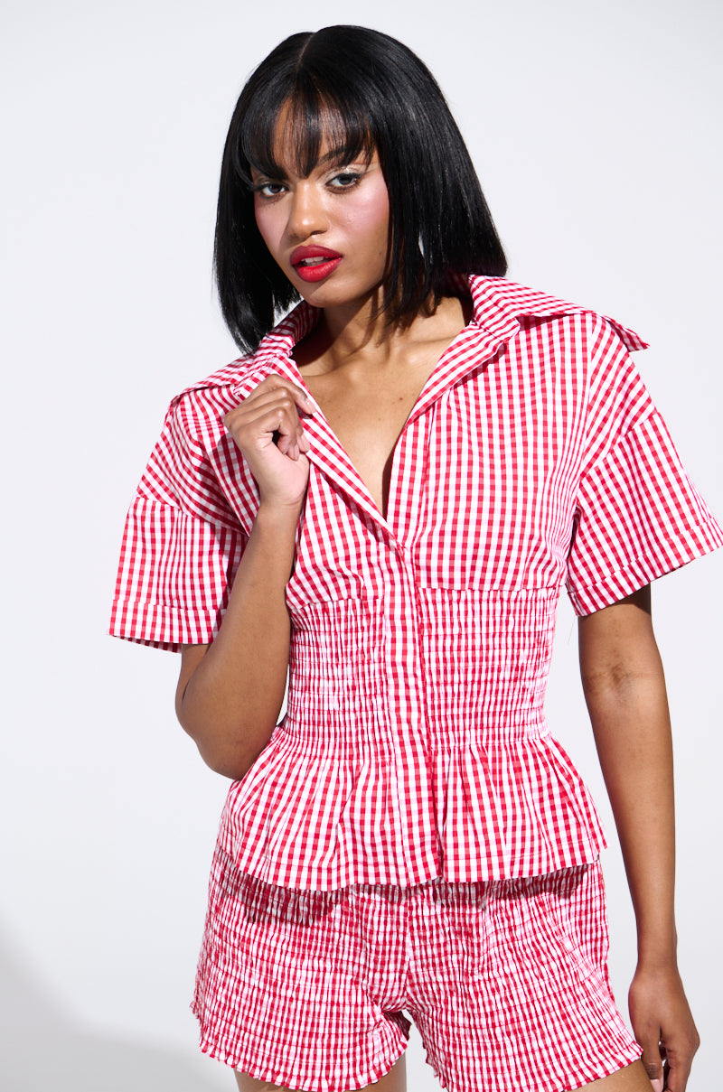 A person confidently poses in a red and white gingham outfit, styled with a PICNIC TIME RUFFLE TOP and matching shorts. With straight black hair and bangs, they face the camera against a plain, light background. 0