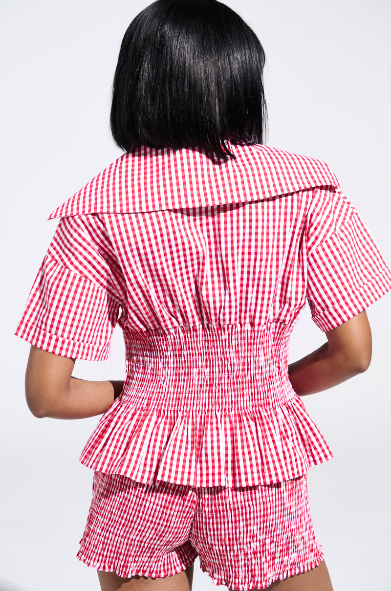 A person is pictured from behind wearing the Picnic Time Ruffle Top, showcasing a red and white gingham pattern with short sleeves, a wide collar, and a seersucker waist. The matching shorts complete the set, all against a plain white background. 2