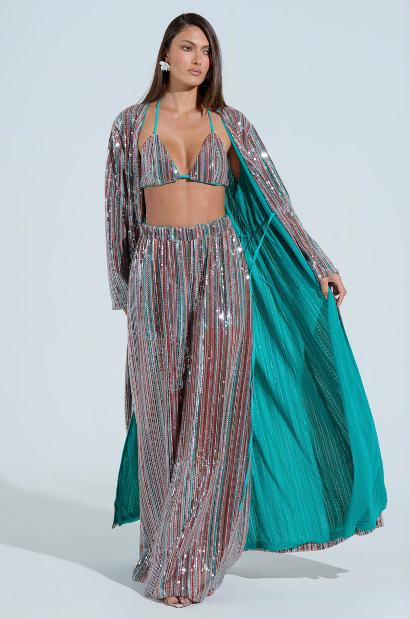 A model showcases a sequin bikini top, AKIRA Label TAYLOR SEQUIN WIDE LEG PANTS, and a matching open-front cape with vertical stripes and bright blue lining, accessorized with large hoop earrings.