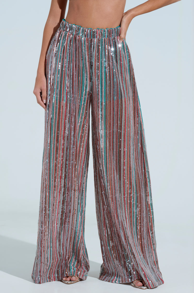 A person wearing the TAYLOR SEQUIN WIDE LEG PANTS captivates with multicolored vertical sequins on a plain background. One hand rests confidently on their hip, as their toes elegantly peek from below the hem.