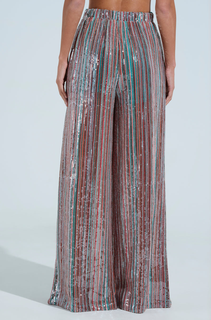 The back view shows a person wearing TAYLOR SEQUIN WIDE LEG PANTS, high-waisted with vertical metallic stripes in silver, green, blue, and red over a light background featuring a chic high-rise design.