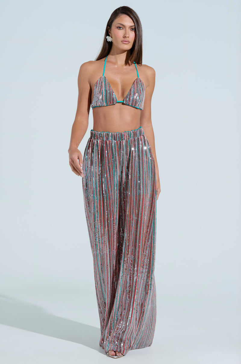 Against a light blue backdrop, a woman dazzles in a colorful, shimmering AKIRA Label halter bikini top and TAYLOR SEQUIN WIDE LEG PANTS. Her cascading hair and statement earrings enhance her captivating look.