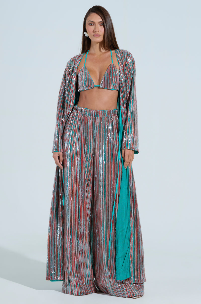 A woman in shimmering, multicolored striped attire featuring a deep neckline and AKIRA Label high-rise trousers poses against a light blue backdrop. Her ensemble, dominated by silver and turquoise hues and TAYLOR SEQUIN WIDE LEG PANTS, is beautifully finished with a long flowing robe.