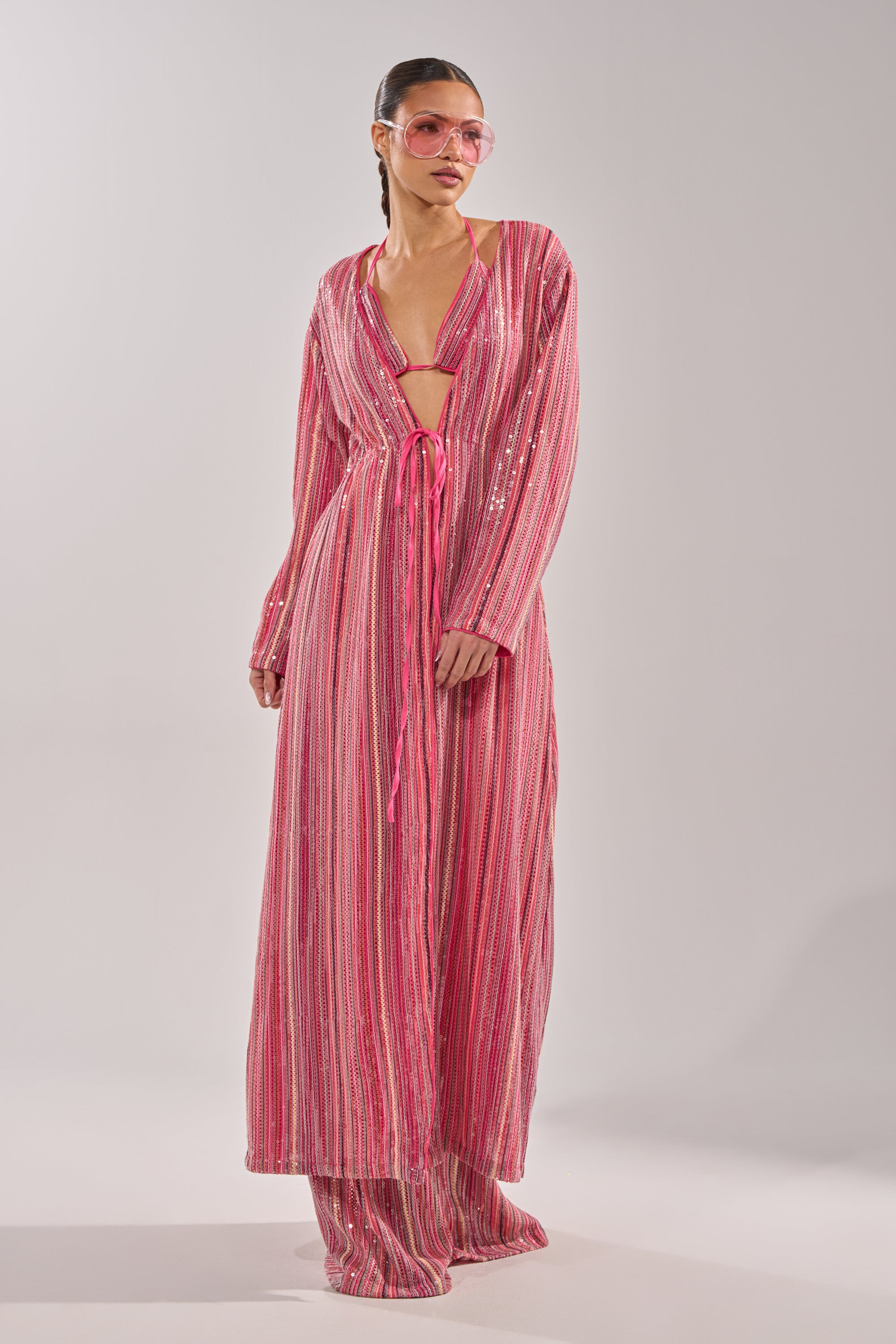 Someone wears the TAYLOR SEQUIN FRONT TIE DUSTER—a long, pink, striped sequin duster with wide sleeves and a waist tie—over matching wide-leg pants and oversized pink sunglasses, creating a standout festival look. 1
