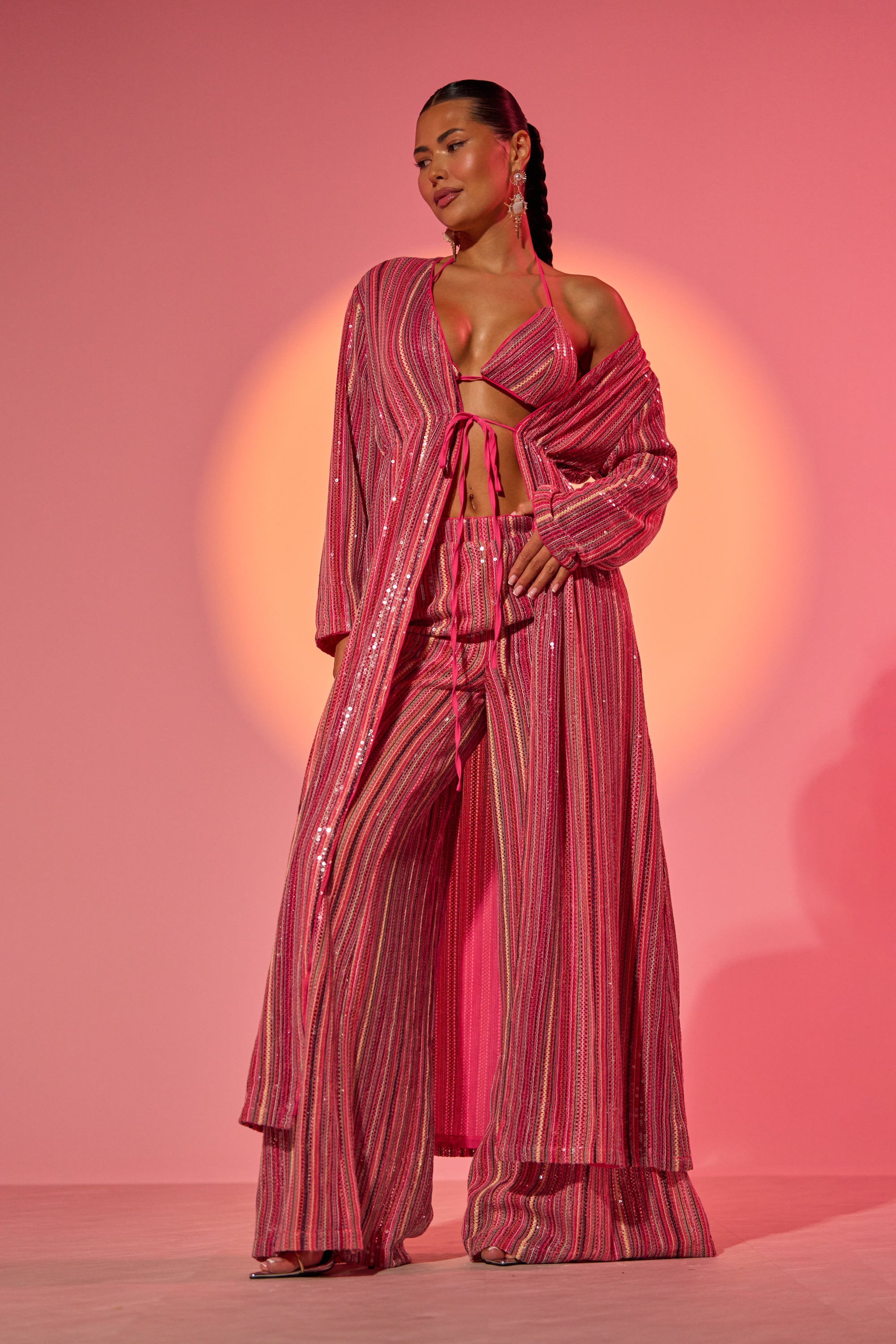 A woman poses confidently in the TAYLOR SEQUIN FRONT TIE DUSTER, paired with wide-legged pants and a bikini-style top, set against a pink backdrop with a glowing circular light behind her. 0