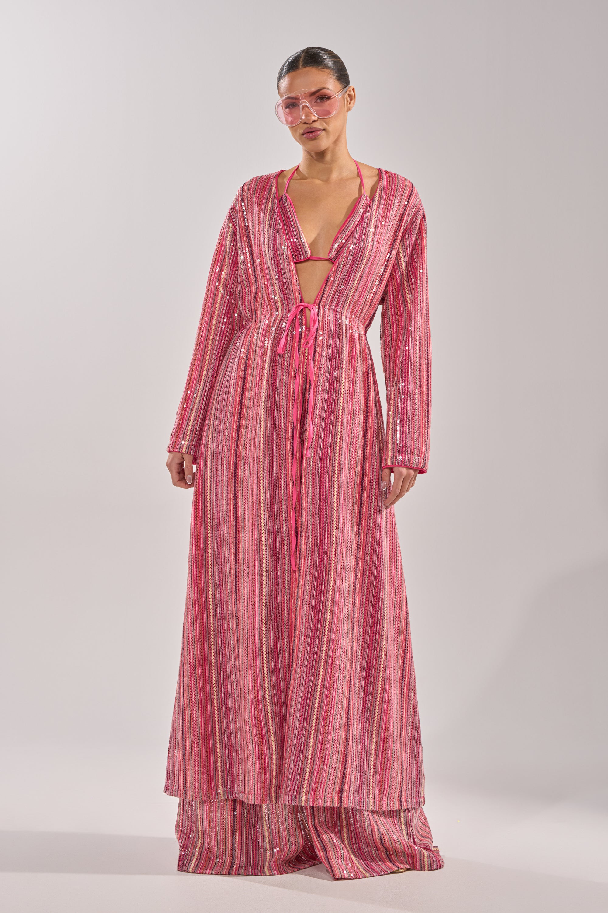 Wearing the TAYLOR SEQUIN FRONT TIE DUSTER over wide-leg pants and pink sunglasses, a person poses against a plain light background, creating a fun festival outfit idea. 5