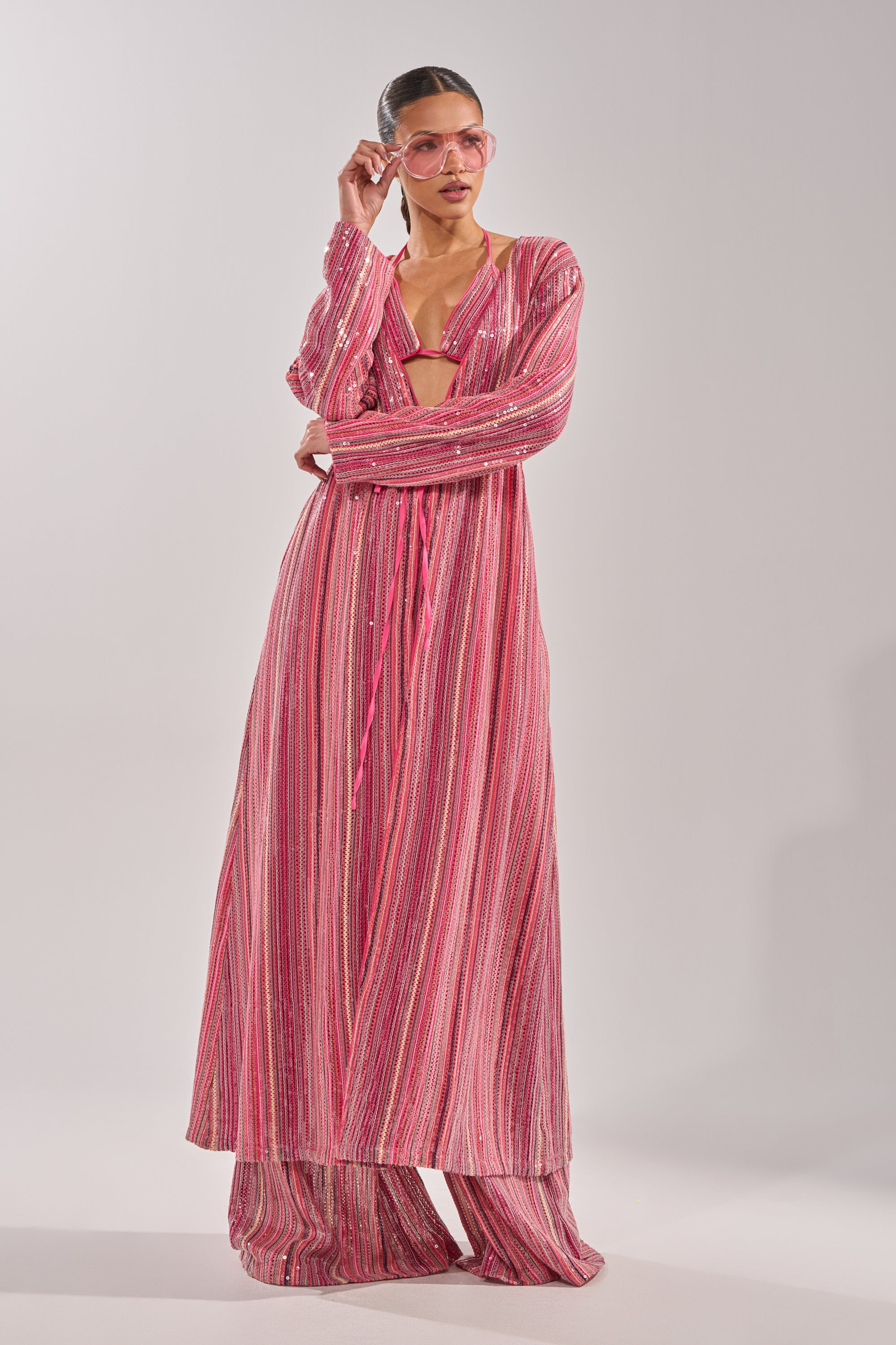 A woman models the TAYLOR SEQUIN FRONT TIE DUSTER, an oversized pink and silver striped festival dress, pairing it with bold pink sunglasses and posing confidently against a plain light background. 6