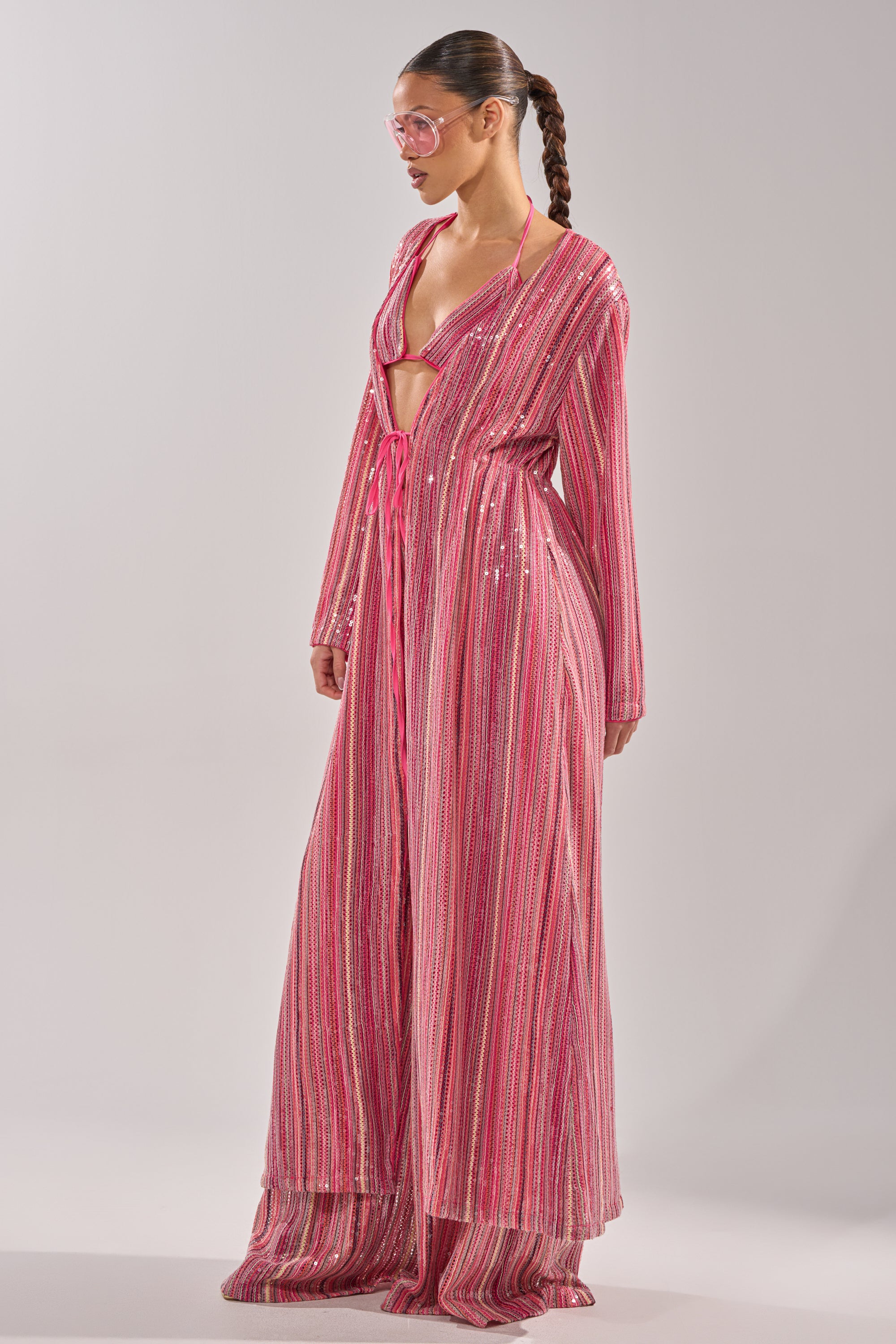 A woman with a braided ponytail wears the TAYLOR SEQUIN FRONT TIE DUSTER over a matching bikini, styled with pink sunglasses for a standout festival look against a plain background. 7