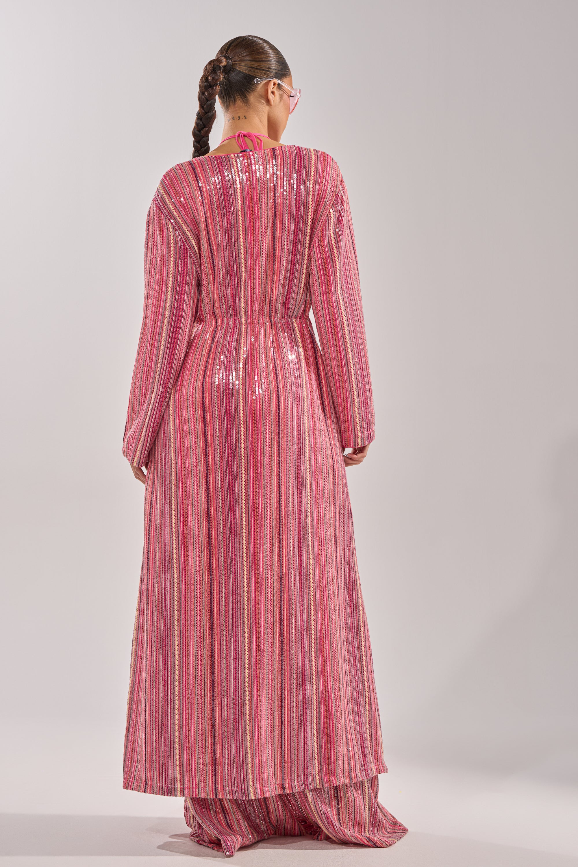 A person with a long braid and pink sunglasses, wearing the TAYLOR SEQUIN FRONT TIE DUSTER—an iridescent pink and silver sequined festival piece—faces away against a light grey background. 8
