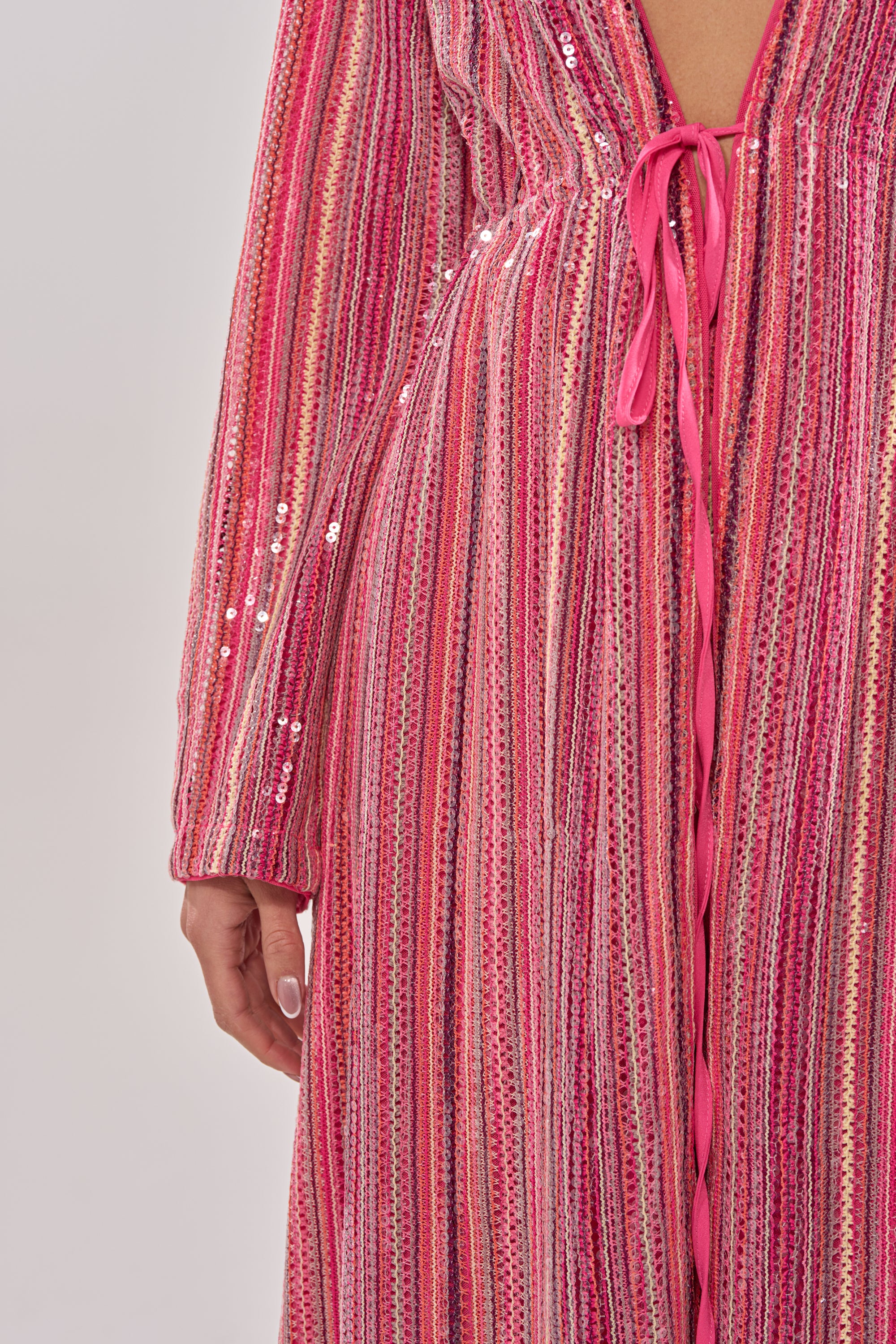 A person wears the TAYLOR SEQUIN FRONT TIE DUSTER—an iridescent, long-sleeved duster with sequins, a deep V neckline, and a pink front tie—ideal for festivals. Only the lower torso and one hand are visible. 9