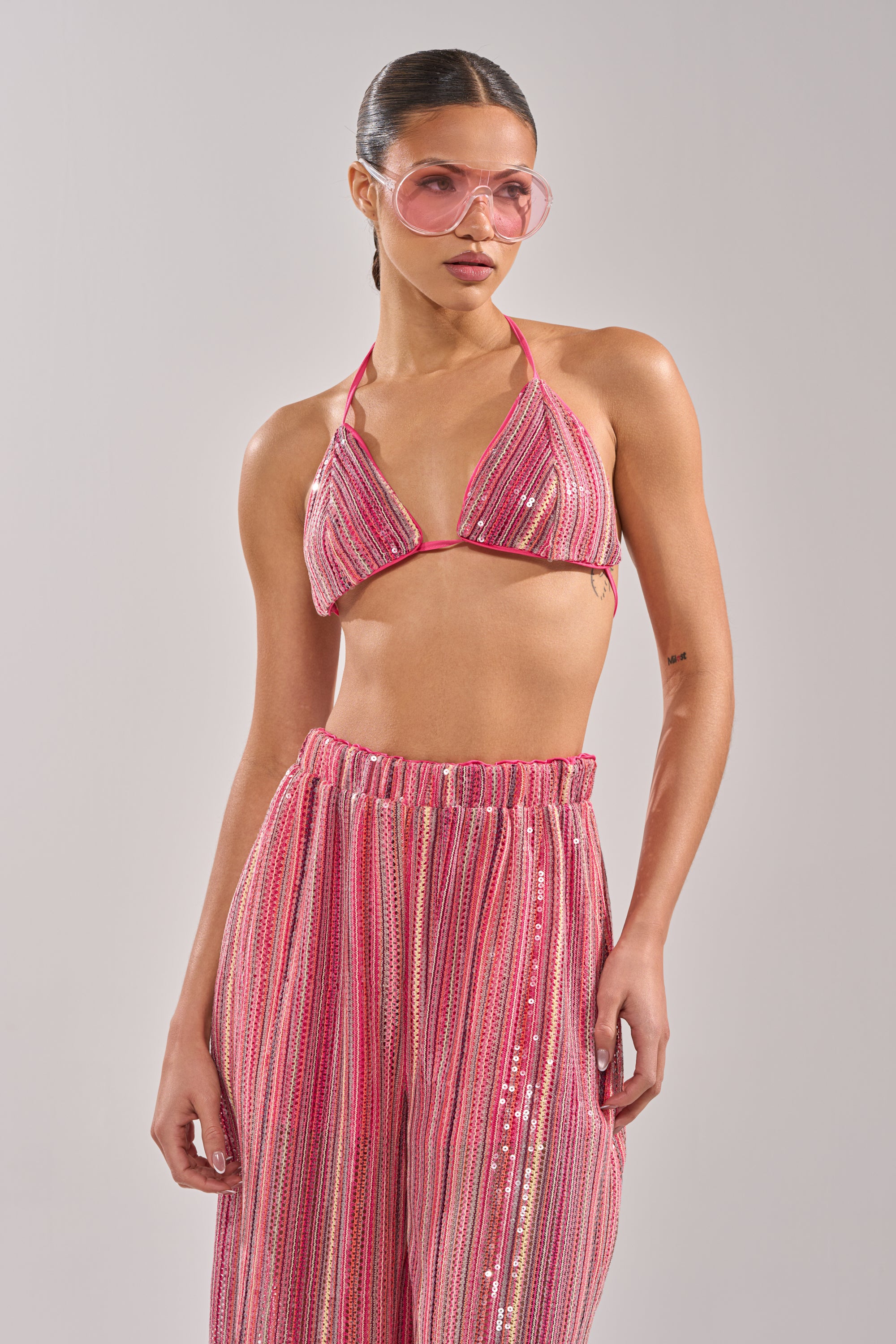 A woman stands against a plain background wearing the TAYLOR SEQUIN BRALETTE, pink-tinted sunglasses, and matching high-waisted wide-leg pants. Her slicked-back hair completes this bold, loungewear-inspired look. 2