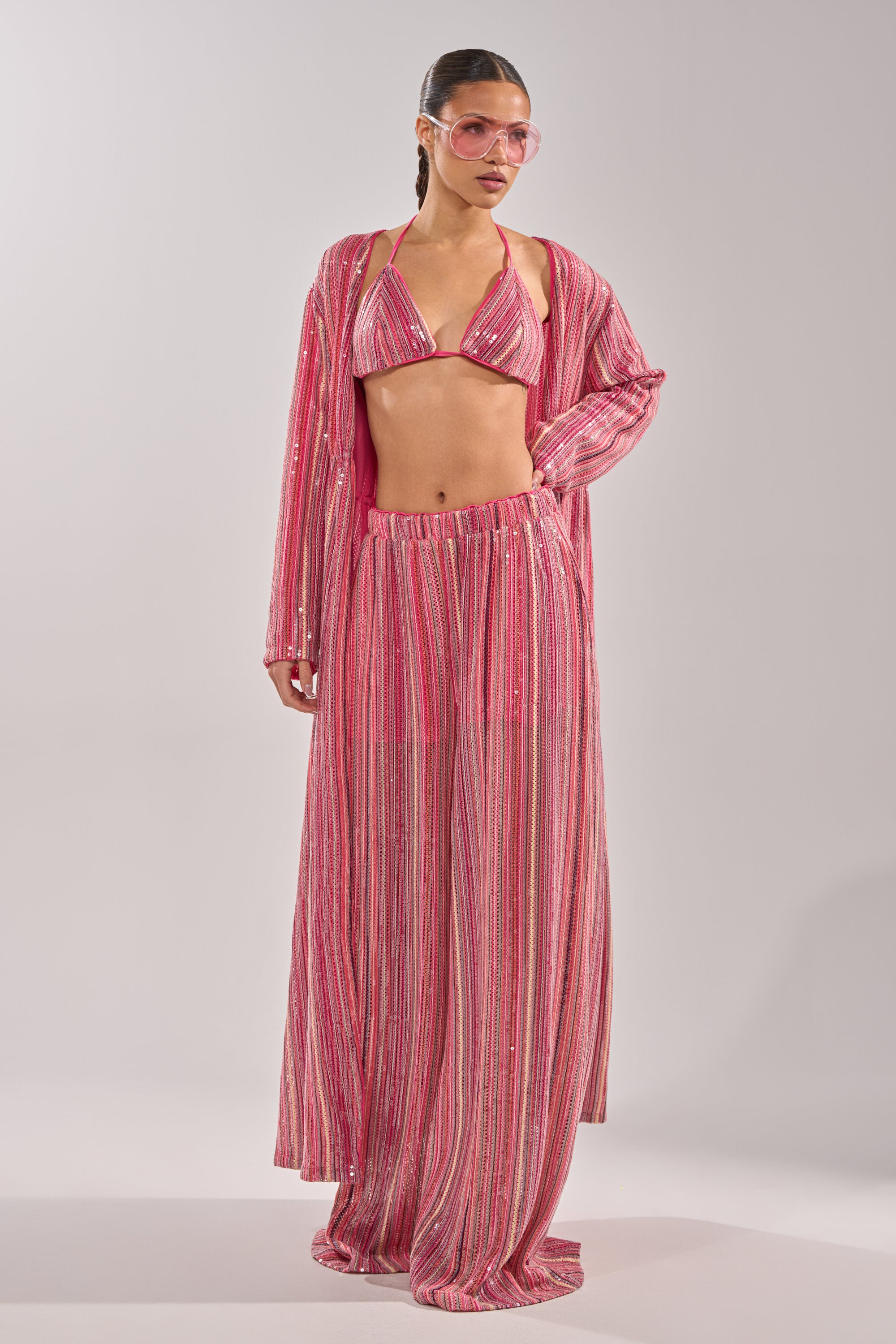 A woman models the TAYLOR SEQUIN BRALETTE in a matching pink, shimmery striped loungewear set with a bikini top, long cardigan, and wide-leg pants. She wears pink sunglasses with slicked-back hair against a plain light background. 11