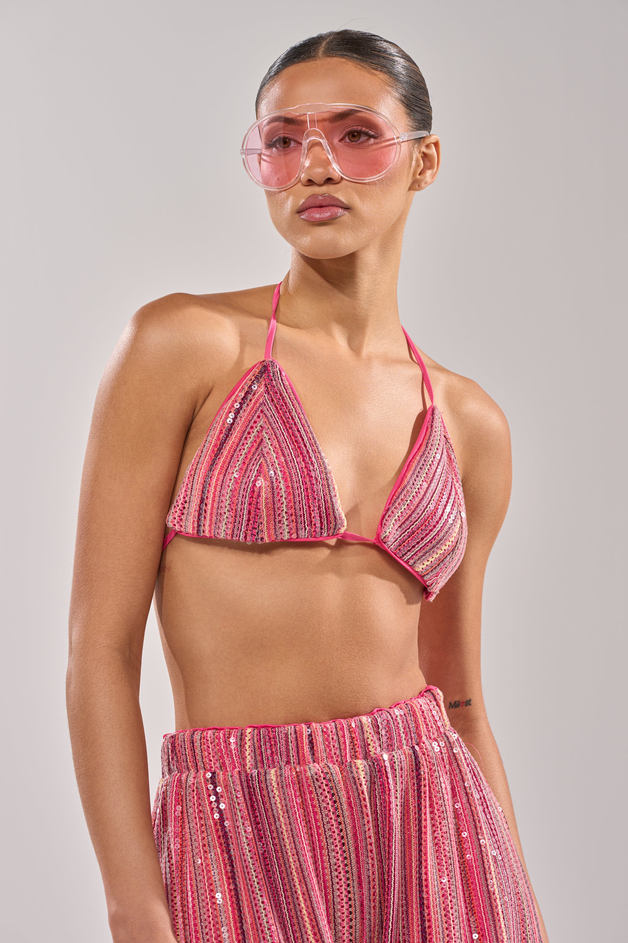 A woman with slicked-back hair and a neutral expression wears pink-tinted sunglasses, the TAYLOR SEQUIN BRALETTE in pink and red stripes, and a matching skirt against a plain background. 3