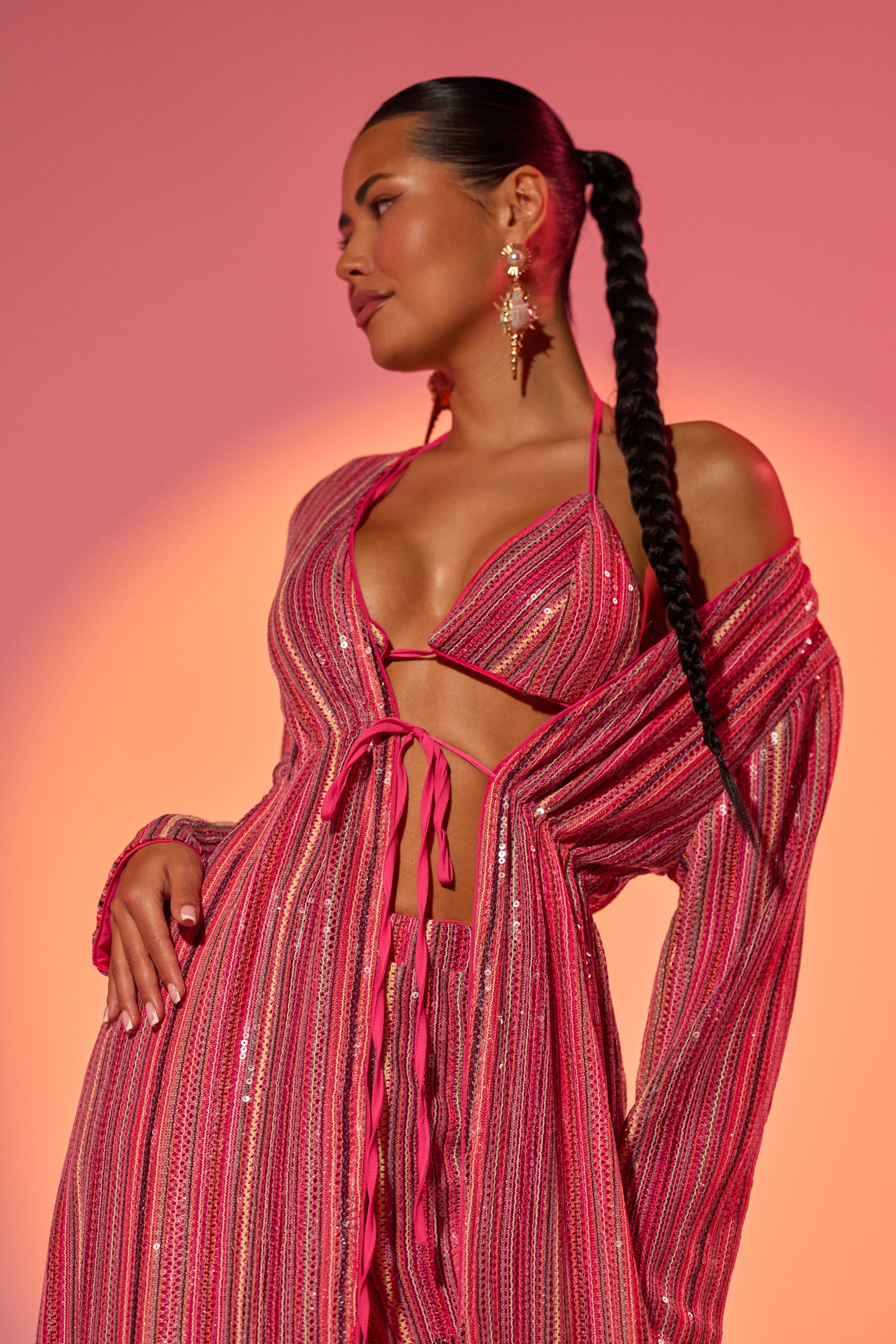 A woman with a long braided ponytail wears the TAYLOR SEQUIN BRALETTE featuring shimmering pink stripes, posing confidently against a warm pink and orange gradient background. 1