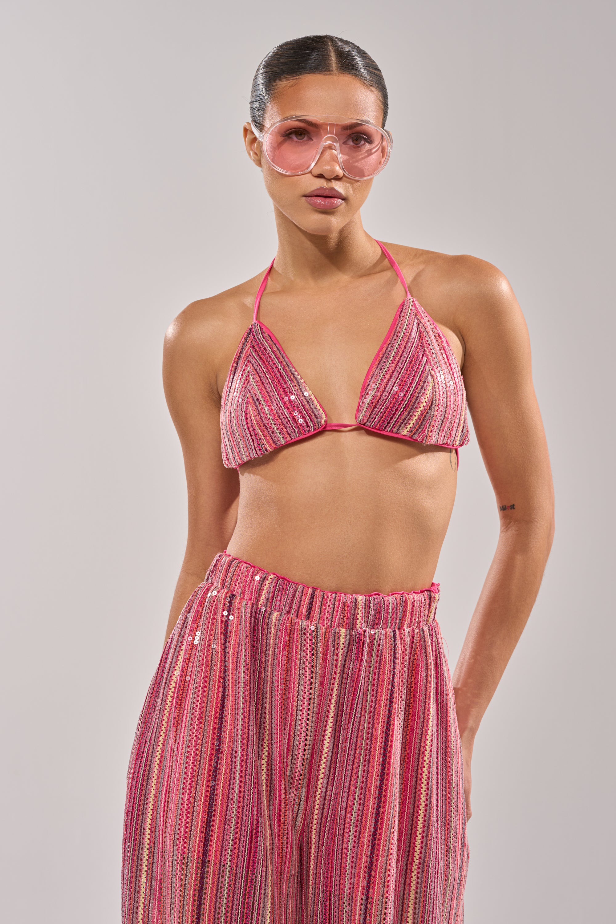 A woman confidently poses against a plain light background, wearing the TAYLOR SEQUIN BRALETTE and matching high-waisted pants, paired with pink-tinted sunglasses. 4