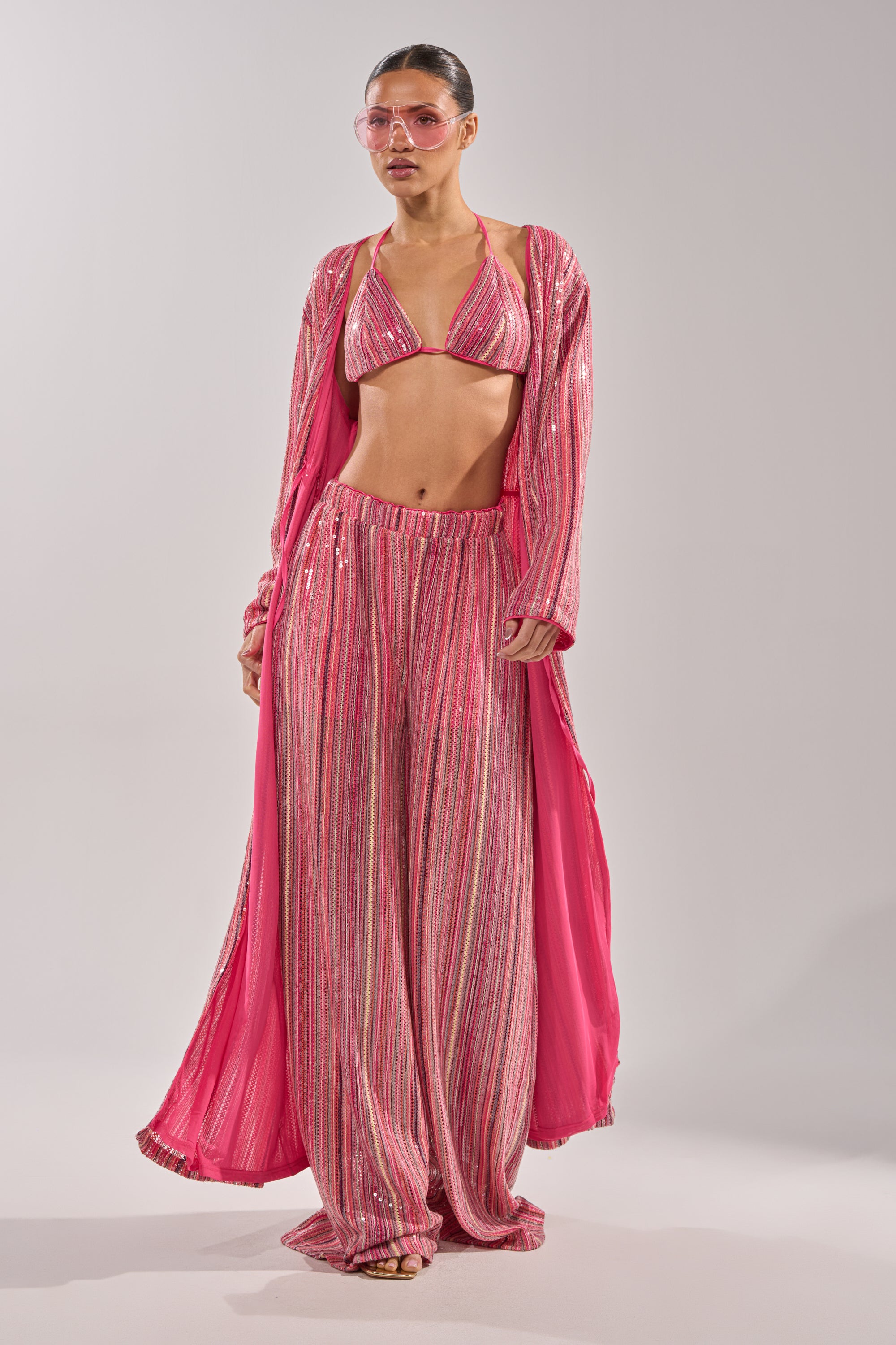 Wearing the TAYLOR SEQUIN BRALETTE, a woman styles a pink, shimmering loungewear set with wide-leg pants and a long duster. She completes her look with pink-tinted sunglasses and slicked-back hair. 9