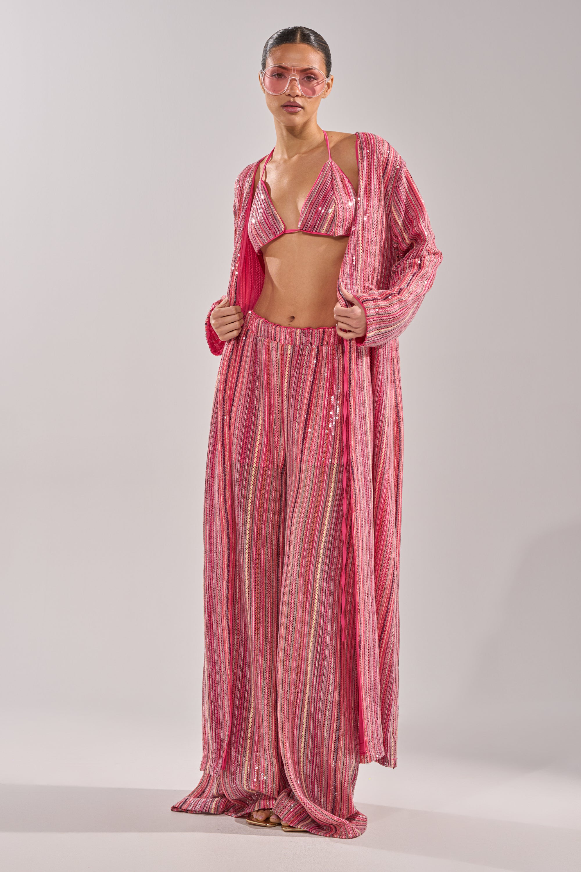 A woman in the TAYLOR SEQUIN BRALETTE bikini top, wide-leg pants, pink-tinted sunglasses, and a matching long, flowy cover-up stands against a plain light background. 10