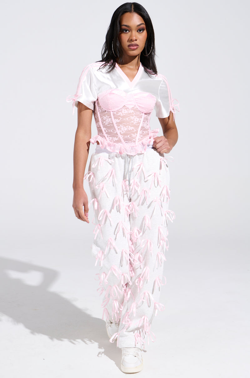 A woman confidently flaunts a white shirt, pink corset, and high-rise white pants with pink satin ribbon appliques. Her long black hair enhances her Girly Pop Jogger style, while white sneakers complete the fashion-forward look against a plain backdrop.