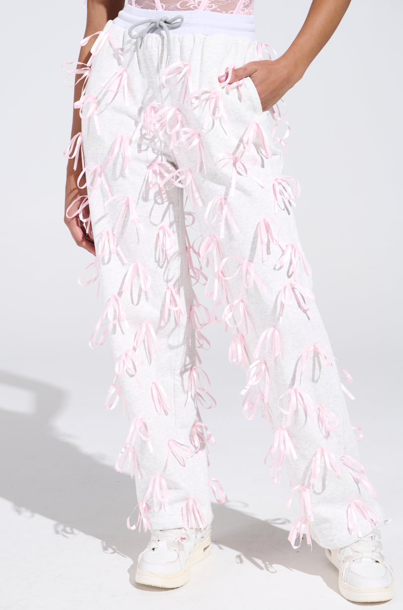 Someone is rocking GIRLY POP JOGGER sweatpants, adorned with satin pink ribbon appliques and a high-rise fit. The pants include a gray drawstring and are complemented by white sneakers, pictured against a simple, light-colored background.