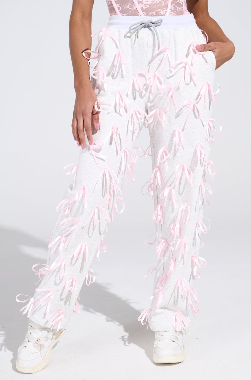 Someone is wearing GIRLY POP JOGGER light gray sweatpants with satin pink ribbon appliques, paired with white sneakers and a cute pink lace top. They beautifully stand out against the plain, soft background.