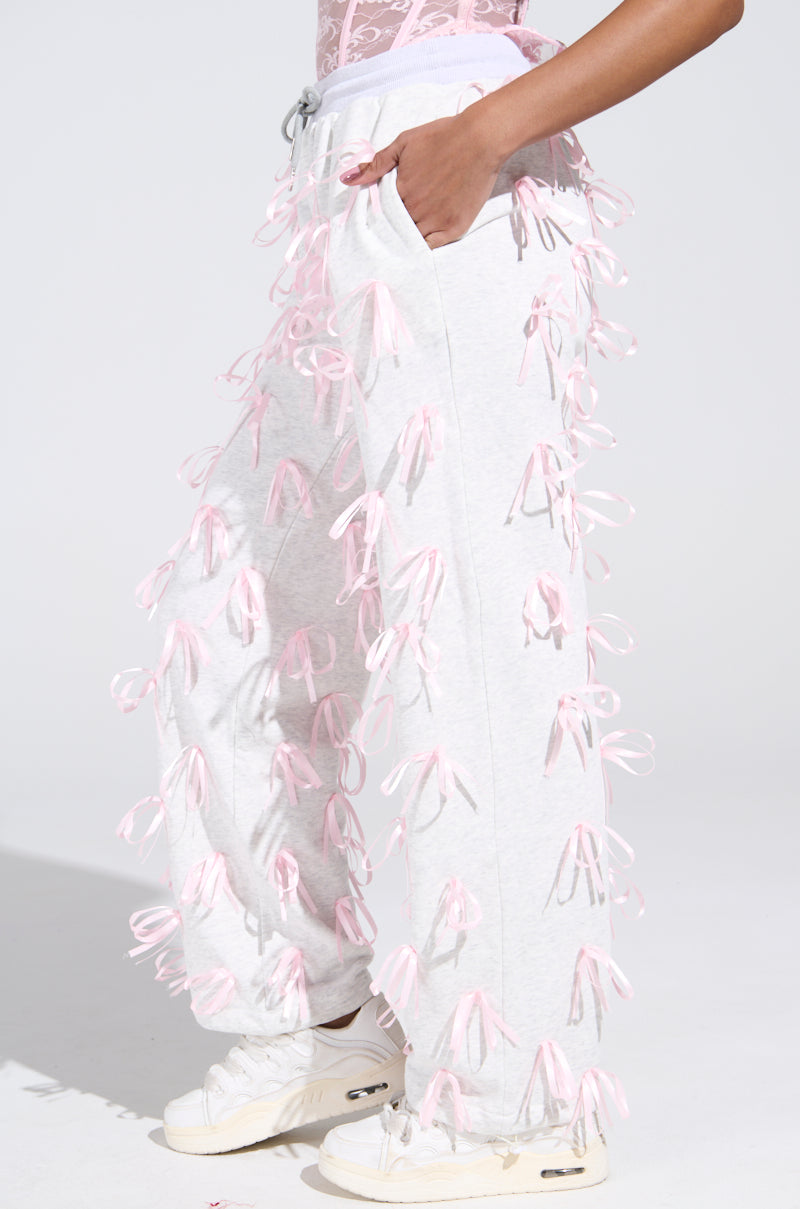 Someone poses against a plain background in GIRLY POP JOGGER pants, featuring white fabric with pink satin ribbon appliques. Paired with a pink lace top and white sneakers, their hand rests in the high-rise fit pocket, highlighting the unique bow detail.