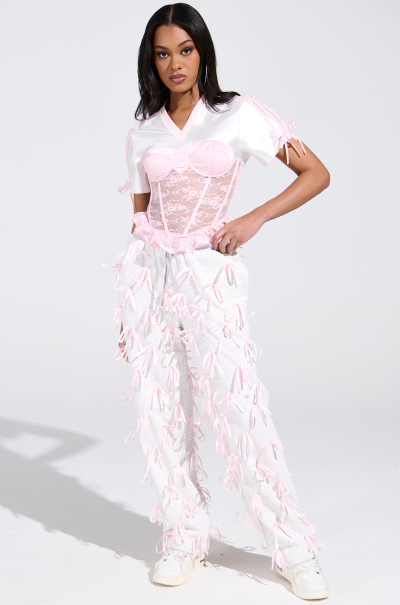 A woman stands confidently against a plain background, wearing the GIRLY POP JERSEY from the AKIRA Label collection. She sports a white top with floral lace corset overlay, white pants with pink bows, and white shoes. Her long dark hair cascades down her back, completing the chic ensemble.