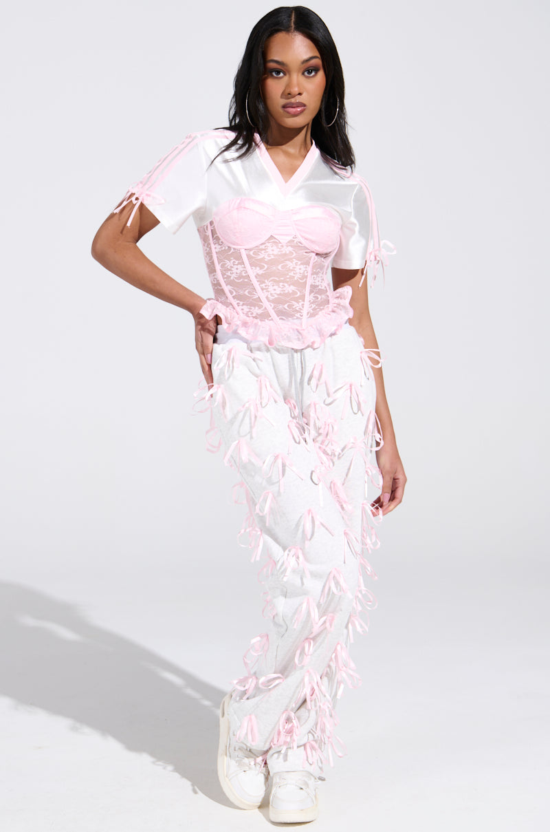 A woman poses against a plain background, showcasing an AKIRA Label GIRLY POP JERSEY look with a white satin top and floral lace corset. Her outfit includes white pants with pink bows, long dark hair, large hoop earrings, and stylish white sneakers.