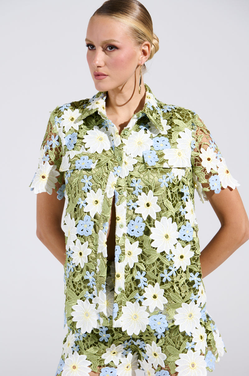 A person models the DAISY CROCHET SHORT SLEEVE BUTTON UP SHIRT, showcasing its embroidered green, white, and light blue floral lace design. They have a sleek hairstyle and wear hoop earrings against a plain background.