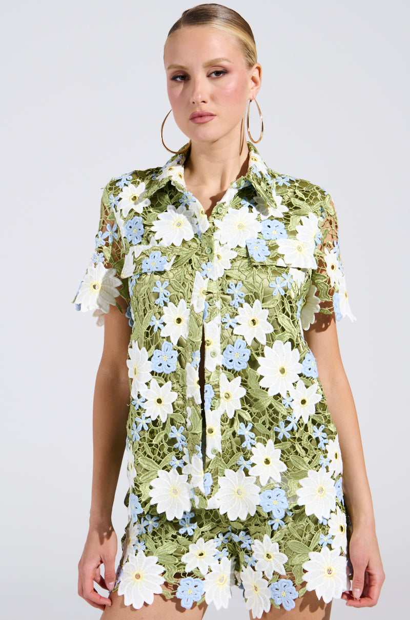 Sporting a sheer green outfit adorned with large white and small blue floral patterns, this ensemble epitomizes Spring Fashion. The DAISY CROCHET SHORT SLEEVE BUTTON UP SHIRT pairs with matching shorts, hoop earrings, and pulled-back hair against a plain backdrop.