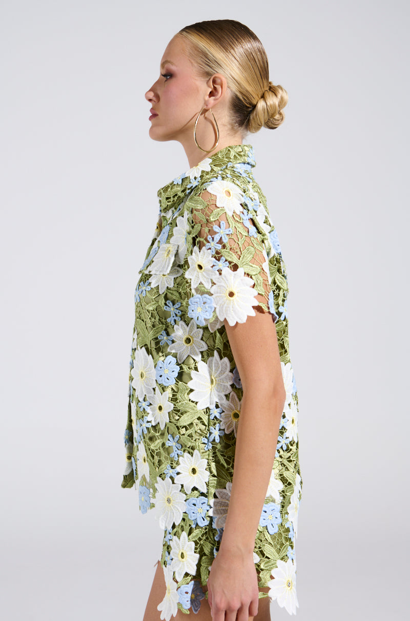 A woman in profile wears a DAISY CROCHET SHORT SLEEVE BUTTON UP SHIRT, featuring green lace with white and blue florals, a nod to spring style. Her hair is styled in a low bun, complemented by large hoop earrings. She stands against a plain light background, exuding embroidered floral elegance.