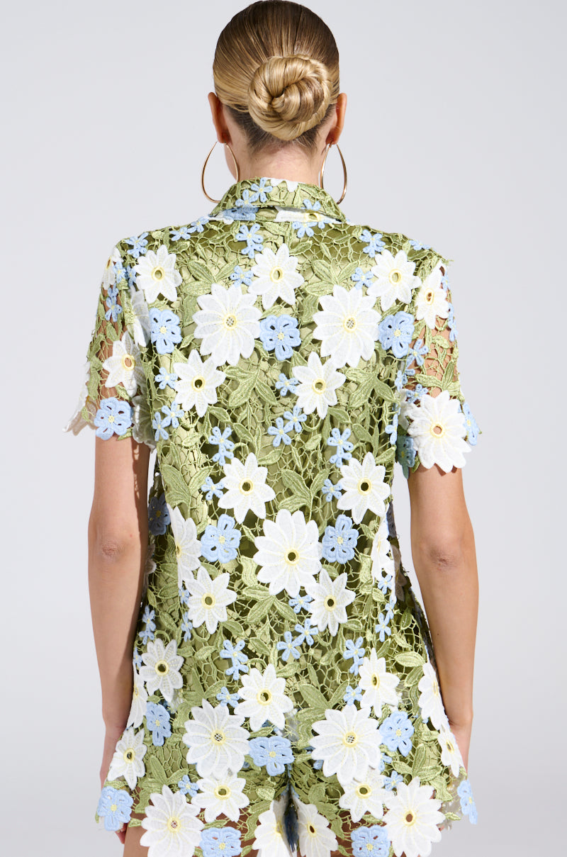 A person with a bun hairstyle wears the DAISY CROCHET SHORT SLEEVE BUTTON UP SHIRT, a floral lace top in green with white and blue flowers. They sport large hoop earrings, capturing spring fashion against a neutral backdrop.