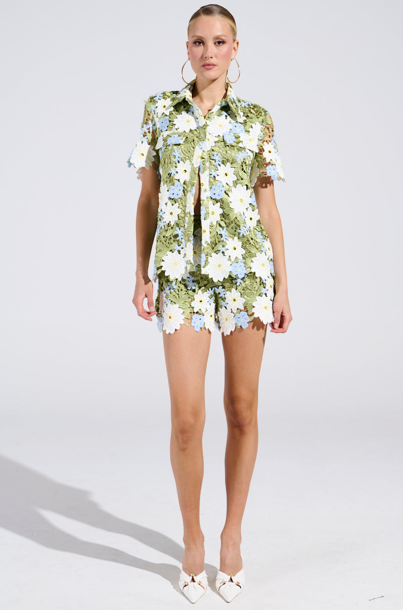 Against a green backdrop with white flowers, a person dons a DAISY CROCHET SHORT SLEEVE BUTTON UP SHIRT and matching shorts, complemented by white pointed shoes and hoop earrings. Their hair is elegantly pulled back, creating a perfect spring fashion ensemble.