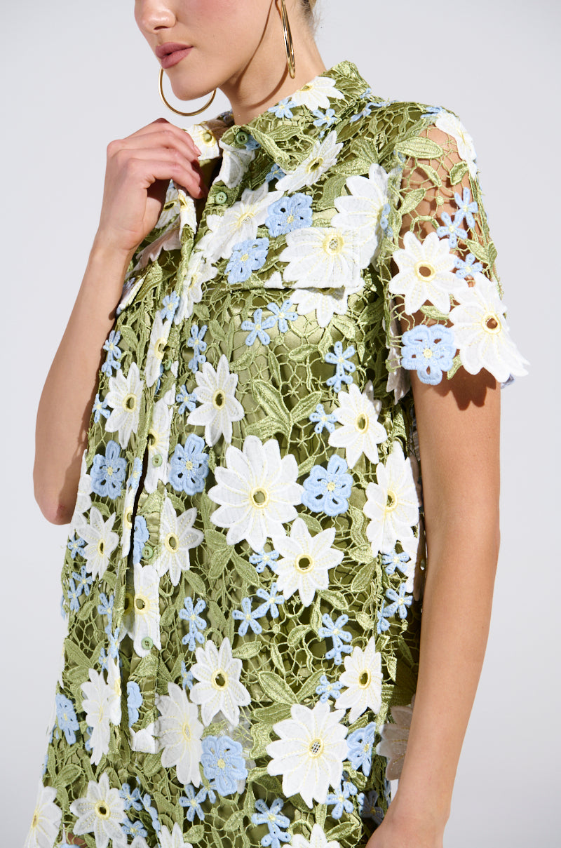 A person dons the Daisy Crochet Short Sleeve Button Up Shirt, a green floral lace piece with elegant white and blue embroidered flowers that capture spring fashion. The shirt features a charming collar, which the individual lightly touches.