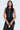 A woman in a sleeveless GIVE ME LOVE BLOUSE with a vest-like silhouette and shorts, adorned with decorative silver bow accents down the front, totes a small black handbag with a satin-lined interior. She pairs it with sheer black tights against a plain white background.