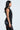 A person with long dark hair stands in profile against a plain background, wearing the sleek GIVE ME LOVE BLOUSE with decorative clasps and black shorts, holding a chic black bag.