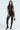 A woman stands wearing a GIVE ME LOVE BLOUSE in a vest-like silhouette with decorative strings, paired with black shorts, black sheer tights, high-heeled shoes, and carrying a small black bag, gazing forward against a plain background.
