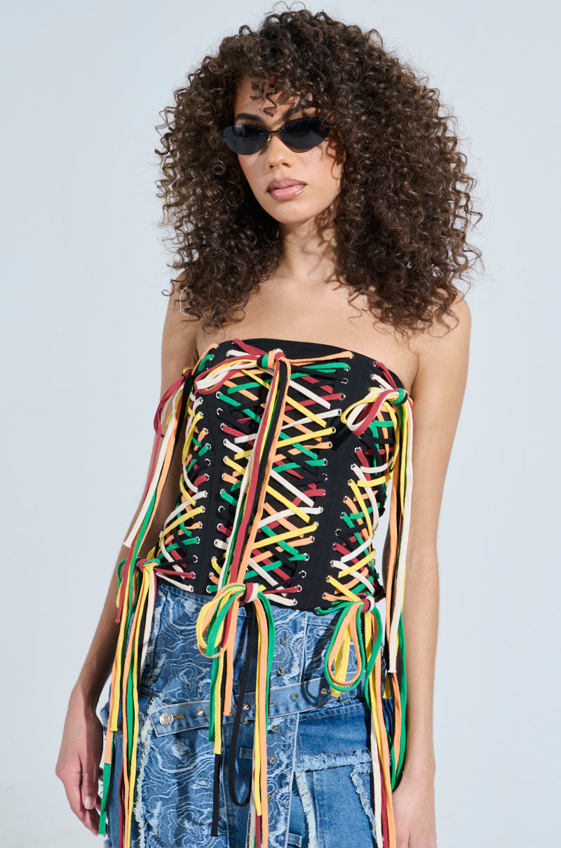 A person with curly hair exudes streetwear vibes, wearing sunglasses and the CROSSED THE LINE CORSET with colorful front crisscross laces. They pair it with patchwork distressed denim against a plain, light-colored backdrop.