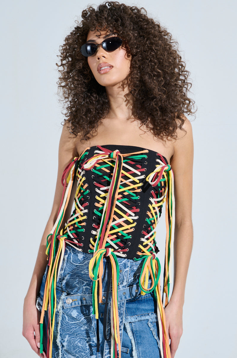 A woman with curly hair rocks the CROSSED THE LINE CORSET, a black strapless waist-snatching top featuring colorful lace-up details in green, yellow, and red. Blue patterned jeans and oval sunglasses complete her streetwear vibe against a plain background.