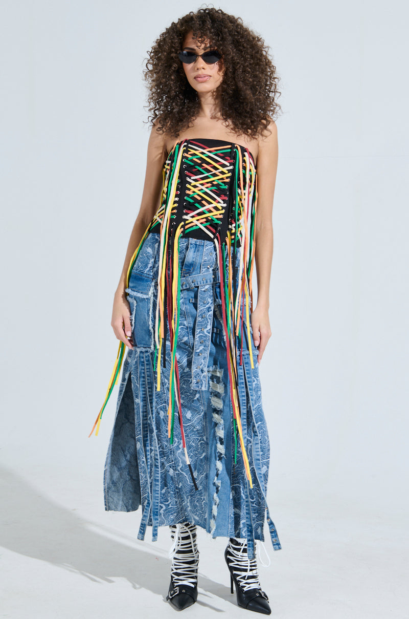 A woman exudes streetwear vibes with her sunglasses, the CROSSED THE LINE corset top accentuating her waist, and a patchwork denim skirt with a front slit. Her curly hair complements the look, finished with black and white strappy shoes against a light background.