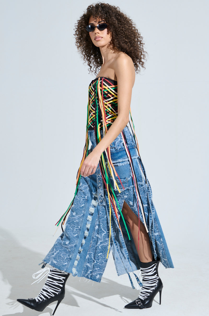 A fashionable woman walks confidently in the CROSSED THE LINE CORSET with colorful strings, paired with a long denim skirt featuring side slits. Completing her modern streetwear look are sunglasses, lace-up shoes, and her curly hair against a plain background.