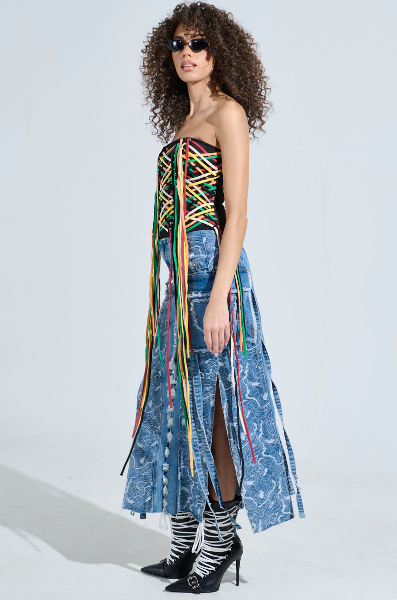 A curly-haired individual sports sunglasses, the CROSSED THE LINE CORSET with colorful strings, and a long blue patterned skirt. Black and white patterned boots complete their look against a plain backdrop.