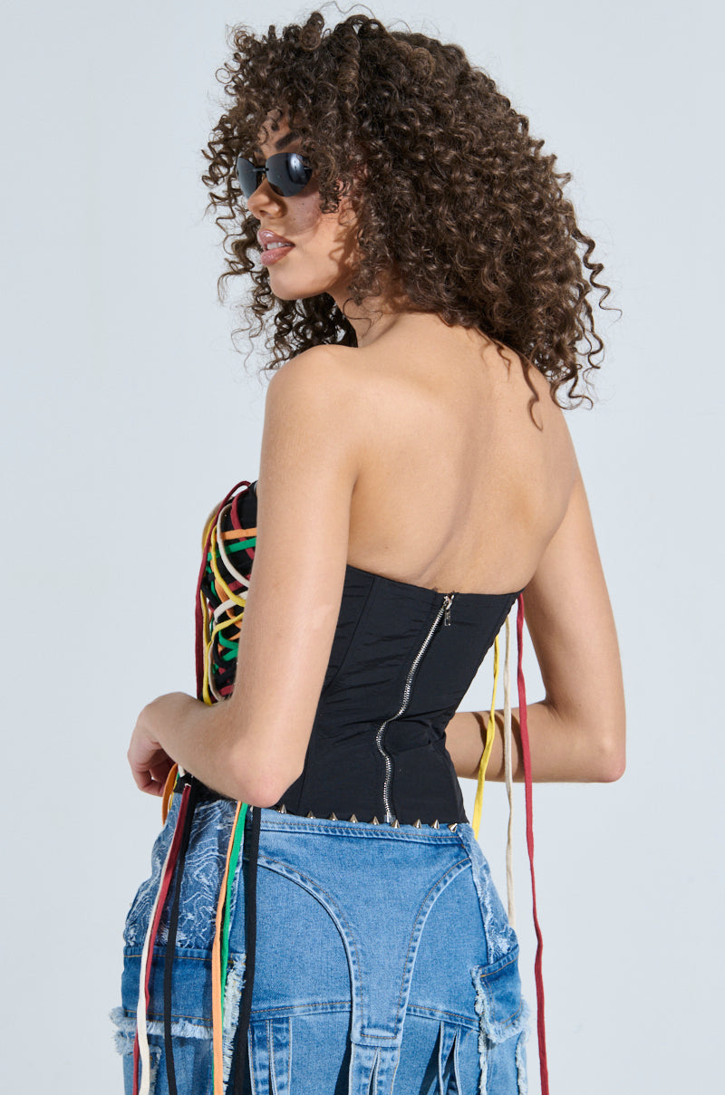A person with curly hair, sunglasses, and a strapless black top adorned with colorful strings stands confidently. The CROSSED THE LINE CORSET, featuring lace-up panels, adds a chic touch around the waist, perfectly complementing the plain, light-colored background.