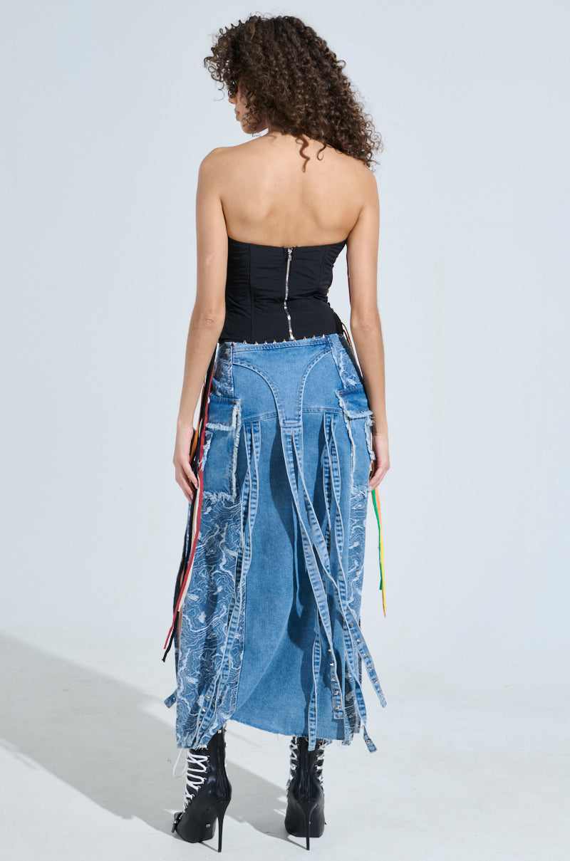 A person with curly hair wears the CROSSED THE LINE CORSET, a strapless, waist-snatching black top, paired with a blue denim skirt featuring fringe and colorful strings. Their look is completed by black lace-up boots as they stand against a plain, light-colored background, facing away from the camera.