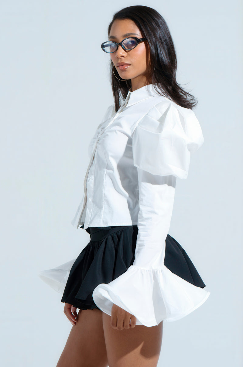 Wearing the stylish DANIELLA BUTTON UP, a cropped white blouse with puffed sleeves, paired with a black skirt and sunglasses, they confidently pose against a light background while glancing to the side. 3