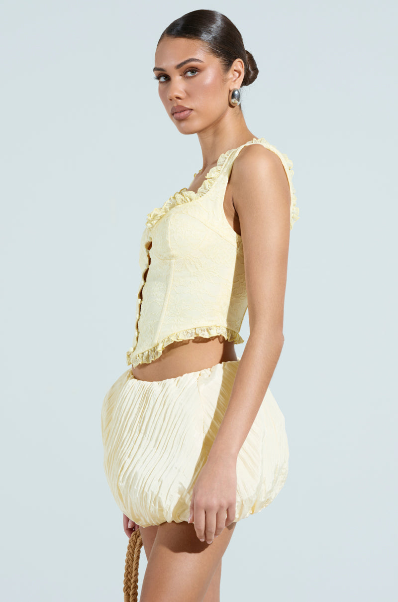 A person wears a SWEETHEART LACE CORSET styled with a balloon skirt. With an updo, they look sideward, highlighting earrings and a satin-lined hint under the ensemble against the plain backdrop.